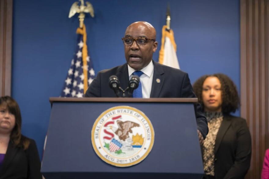 Illinois Attorney General Kwame Raoul stands at a podium with two microphones and a state of Illinois seal -- he's speaking to reporters wearing a black suit with a white shirt, blue tie and Illinois lapel pin.