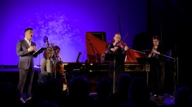 A vocalist sings into microphone accompanied by musicians on viola da gamba, harpsichord, violin and oboe.