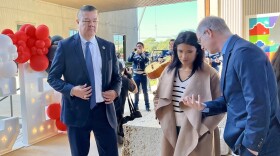 Two men in suits talk to a woman in a tan coat as a mariachi band plays in the background.