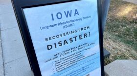 A blue sign is on a sidewalk. It says Iowa Long-term Disaster Recovery Center and "Recovering from Disaster" in black lettering. There is more text on the bottom.