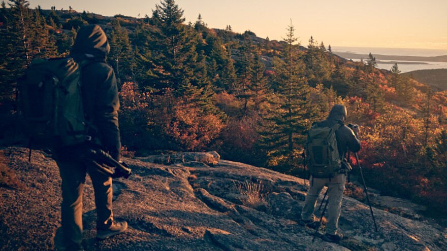 two hikers at the top of a mountain in peak fall, overlooking trees and some water in the far distance