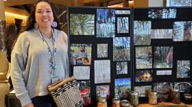 Teara Farrow Ferman stands in front of a display of photographs and containers documenting and displaying first foods. Farrow Ferman wears large, colorful, circular beaded earrings, she has long, dark hair and is smiling.