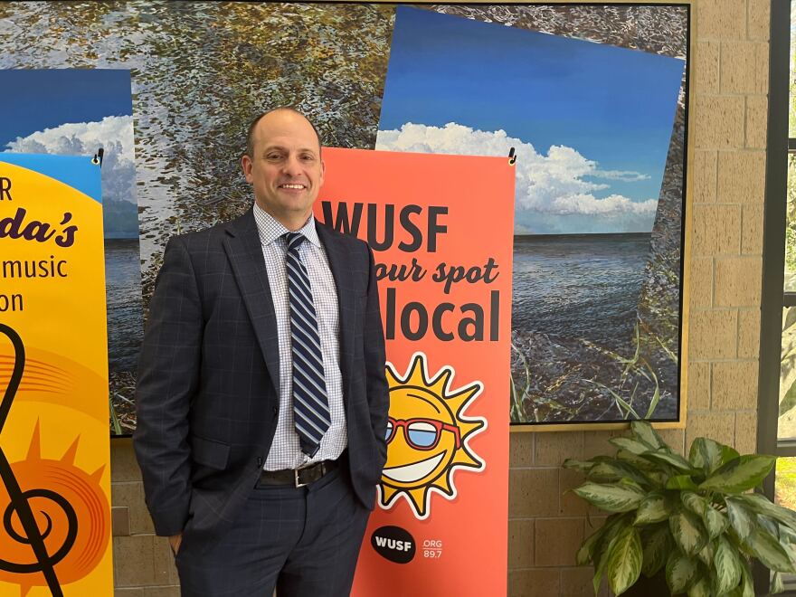 Bald man in suit stands next to WUSF sign smiling 