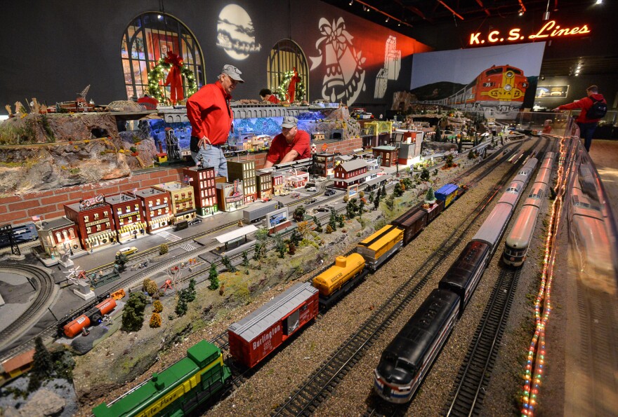 Two men wearing bright red shirts and train conductor caps work amidst a larger display of model trains, tracks and city depictions inside a room decorated for Christmas.