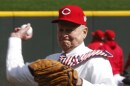 a man in a tie, white button-up shirt, black pants and a cincinnati reds ball cap throws a ball from a pitcher's mound in a baseball field