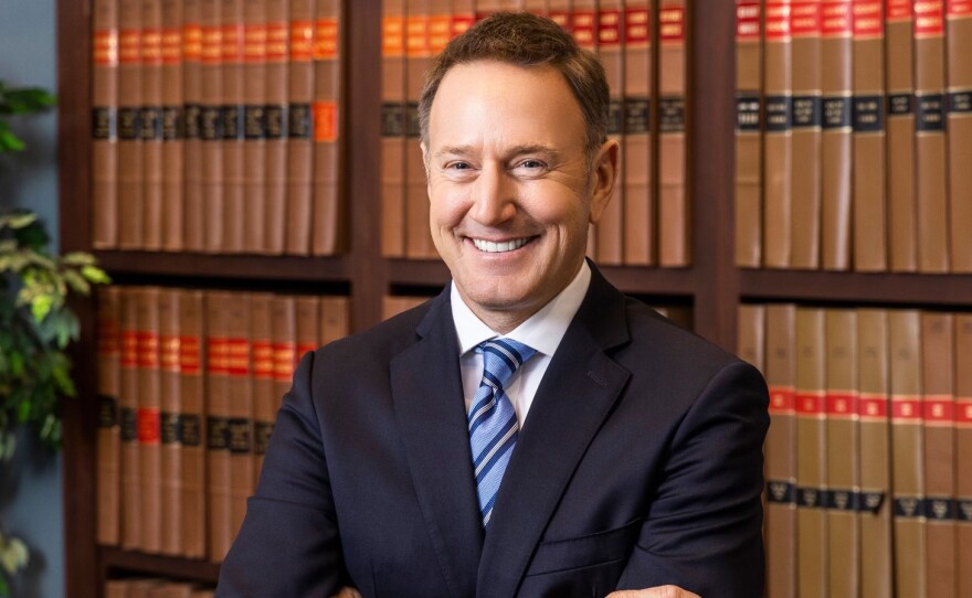 Russell Mock, a white man in a blue suit, stands and smiles in front of shelves filled with books