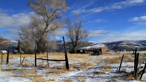 An old, worn-down shed sits in a field with yellow grass and snow. There are old wood fences in the foreground and a mountain range in the background.