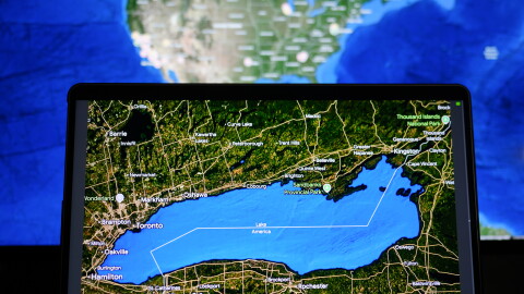 A hand holds up an i-pad that shows a map that is zoomed in on Lake Ontario, which is labeled "Lake America" on the map. Behind it is a computer screen showing a map of the United States. 
