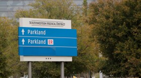 A sign in the Southwest Medical District directing people to Parkland and Parkland's emergency department. The sign is in a median, with several large green trees behind it. The top of the sign, which is grey with black lettering, reads "SOUTHWESTERN MEDICAL DISTRICT." Below that in white block text on a blue background is the words "Parkland" and "Parkland" followed by red block letters in a small white box that says "ER." Both rows have a white arrow pointing up, directing people forward. The third row is also blue, but has been covered up by blue tape to hide what it says.