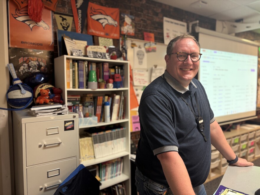 A man in the dark shirt stands in a classroom. 