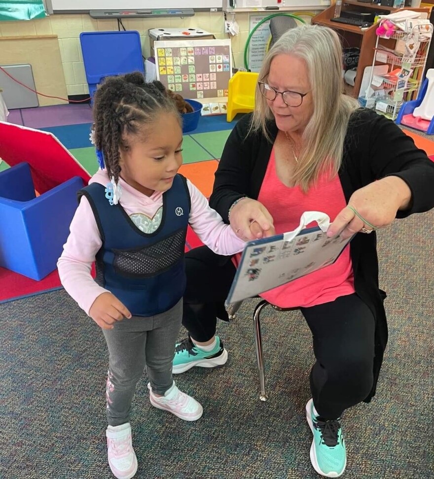 A young child stands next to a seated teacher, who assists the child with pointing at a book. Both are smiling, engrossed in their task.