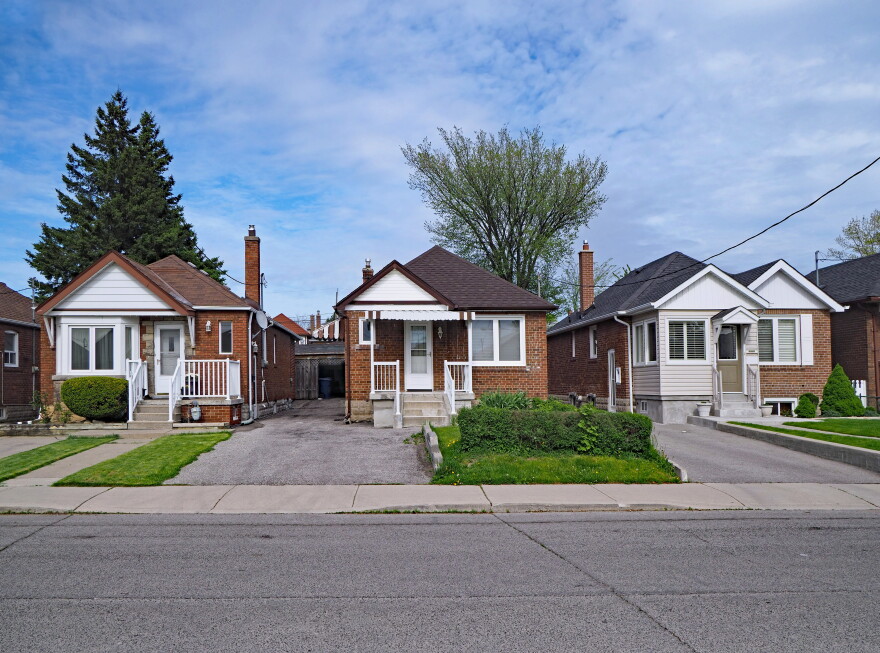 A row of modest brick homes lines a residential street. Each has a small front porch, driveway and neatly trimmed landscaping. The sky is partly cloudy.