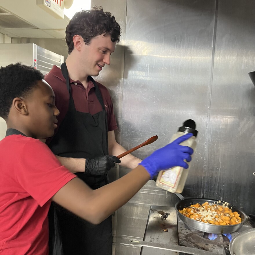 One person pours garlic powder onto sweet potatoes cooking on the stove while another holds the pan by its handle.