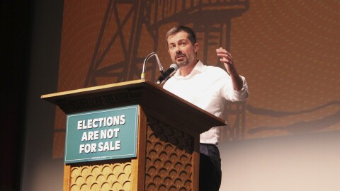 Former Transportation Secretary Pete Buttigieg speaks at a podium inside Butte’s Mother Lode Theater. A sign on the podium reads, “Elections Are Not For Sale.”