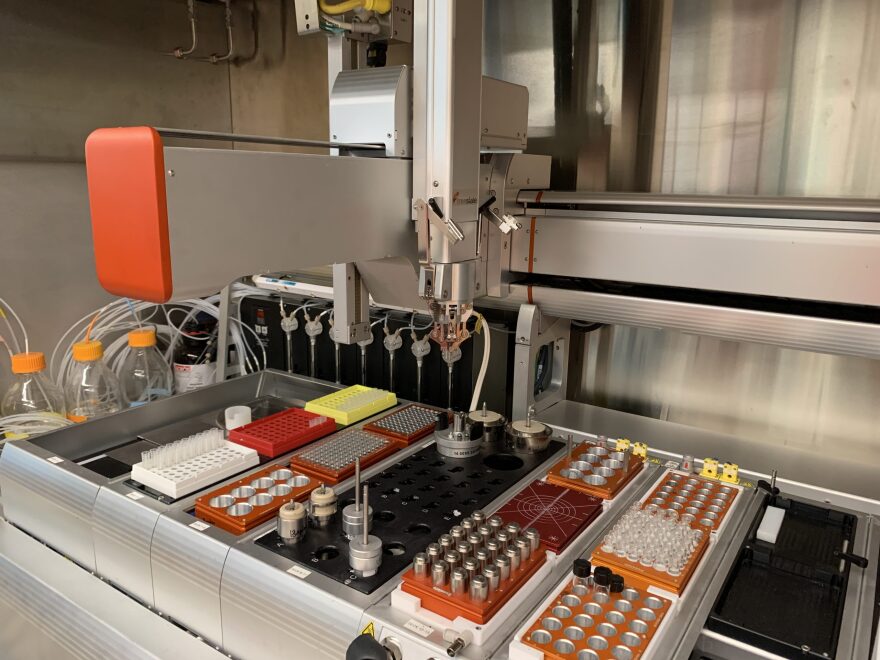 A table of dozens of test tubes sits under a large machine with a mechanical claw used for grabbing the tubes facing down.