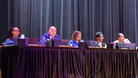 Five adults in suits sit in a line on an auditorium stage at a table with a black curtain in the background.
