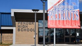 An exterior shot of Pender High School on a sunny morning. The building has a brown brick facade with large windows and a gray concrete roof. On the left side, there are black metal letters reading "Pender High School." On the right side of the photo, there is a large red, white and blue banner bearing the words "Pender Patriots."