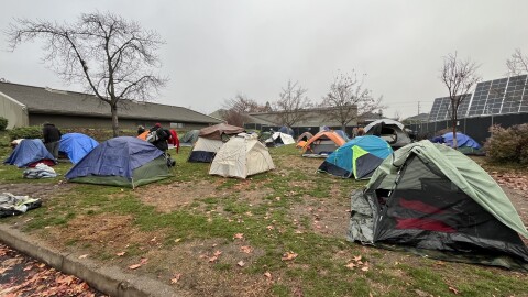 Colorful tents and tarps are set up on a grassy lot. The area is surrounded by buildings and solar panels. 
