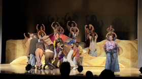 A man wears a colorful long coat that other actors hold as they circle him. Dancers and a narrator stand on stage singing.