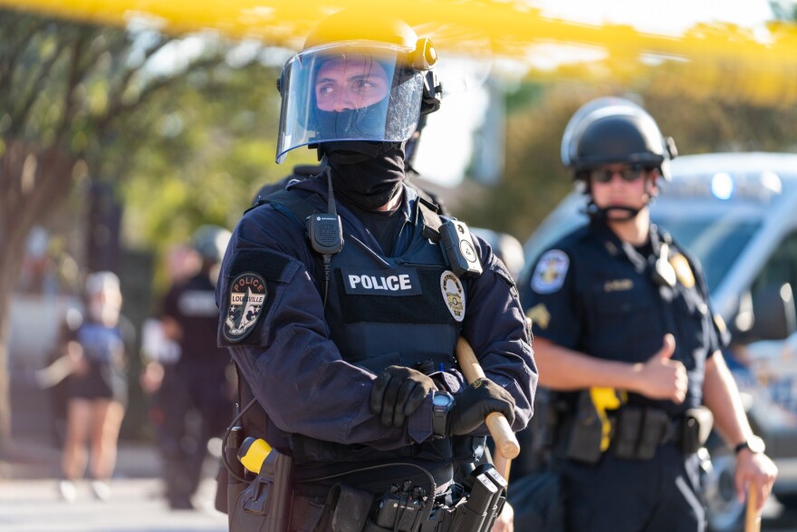 Two police officers wearing helmets, one wearing a face shield, stand, looking away from the camera. A police vehicle is visible behind them. 