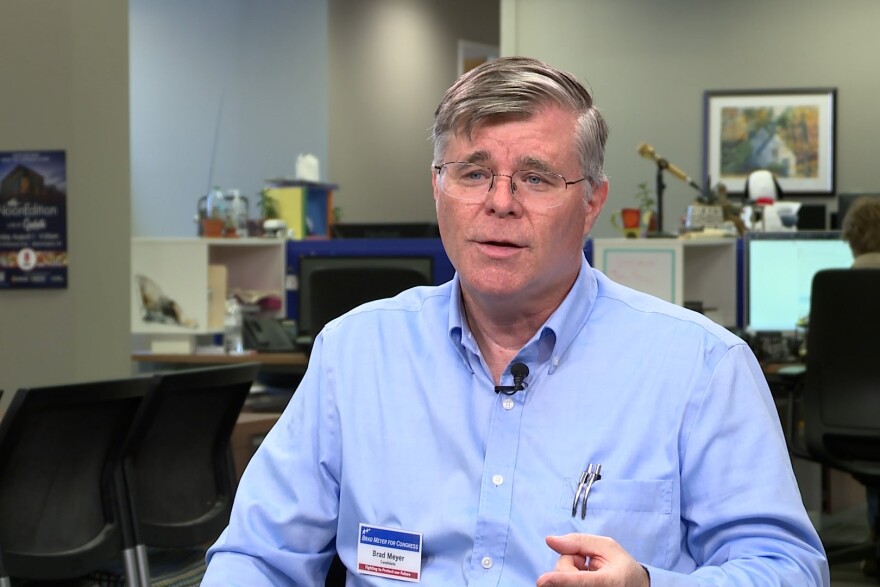 Brad Meyer sitting in front of a camera. He's wearing a blue button down shirt and wire frame glasses. He has gray hair and two pens in his shirt pocket.