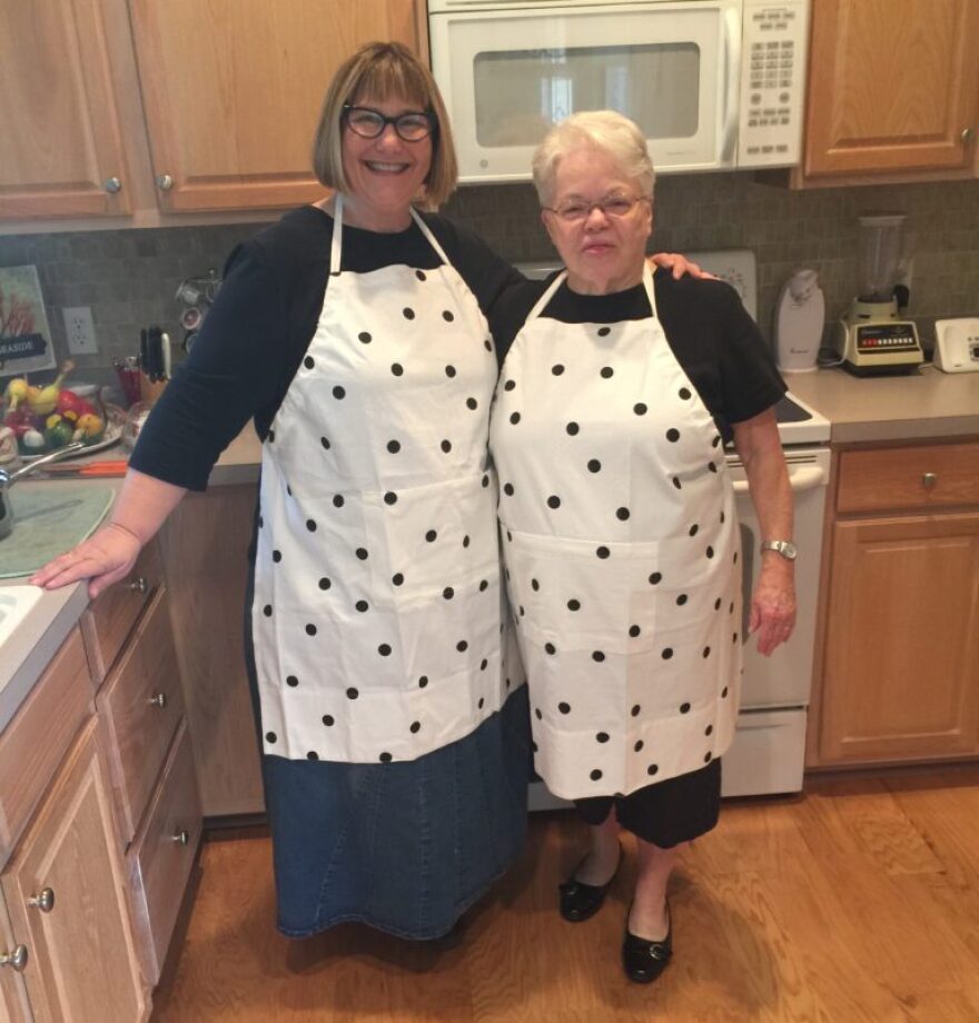 Two women stand together wearing aprons in a kitchen. 