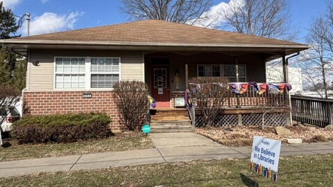 A small house is decorated with rainbow flags. A sign in front reads, "We Believe in Libraries."