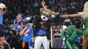 The deer mascot of a professional basketball team poses with its arms crossed at center court of an arena while surrounded by dancers in flamboyant costumes.