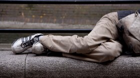 A person in khaki pants and sneakers lies curled up on a stone bench.