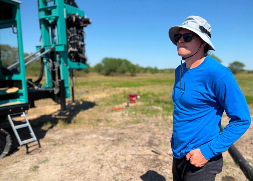 A man in a blue long sleeved shirt, black pants, sun hat and sunglasses is looking thoughtfully into the distance at a piece of heavy machinery.