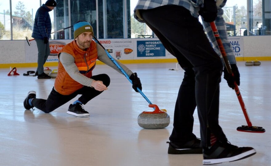 Man slides a curling stone on an ice rink while holding a broom.