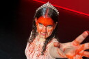 Young woman in pink dress wearing crown covered in fake blood looks up to the camera reaching her bloody hand up 