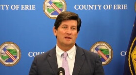 Erie County Executive Mark Poloncarz appears before a microphone in a gray suit, white shirt and lavender tie before a backdrop with the Erie County seal on it