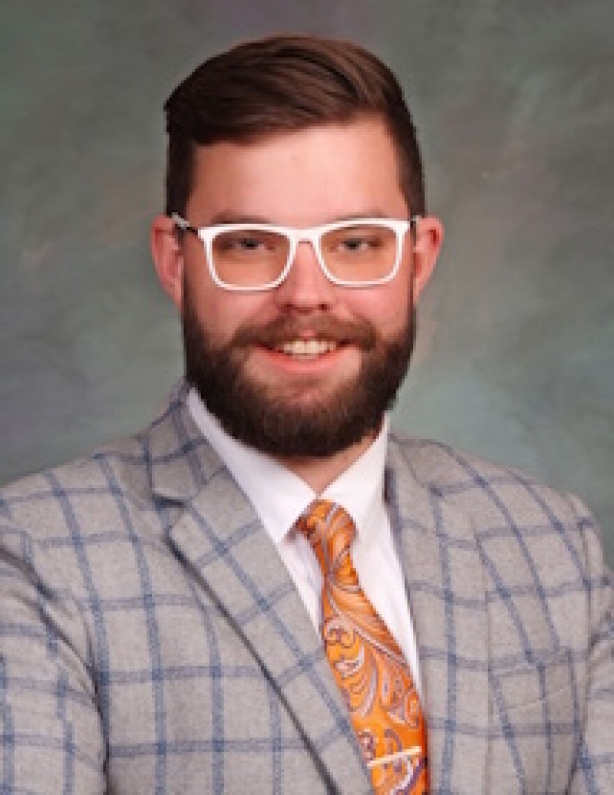 A headshot of a bearded man wearing white rimmed glasses, a light gray suit and orange tie.
