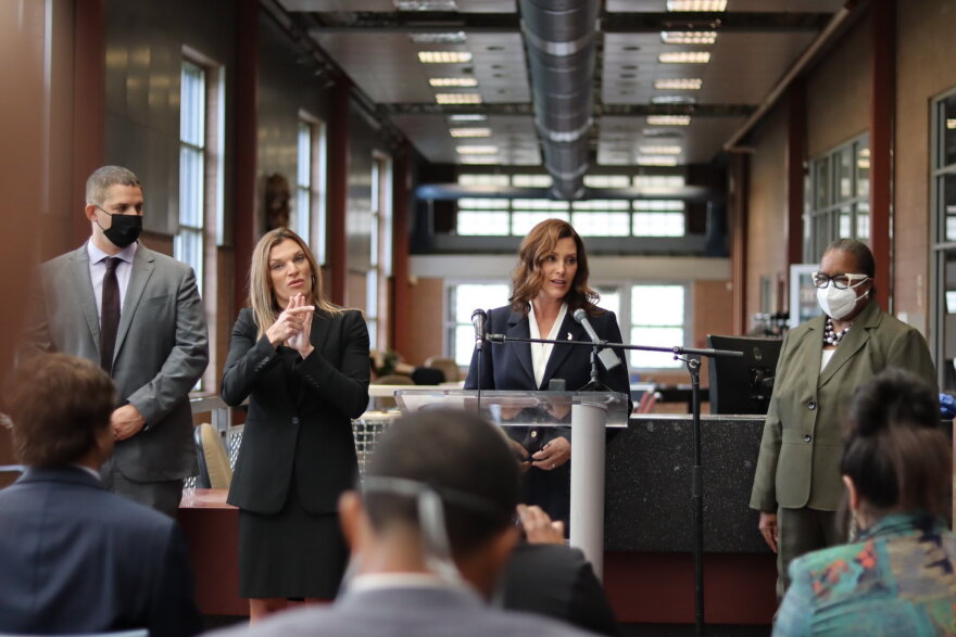 Governor Gretchen Whitmer speaking in front of a crowd at a community center. She speaks at a lectern with two microphone in front of her. There is one woman on her left, our right, wearing a mask facing the crowd. On the other side is Whitmer's American Sign Language interpreter and a man wearing a mask.