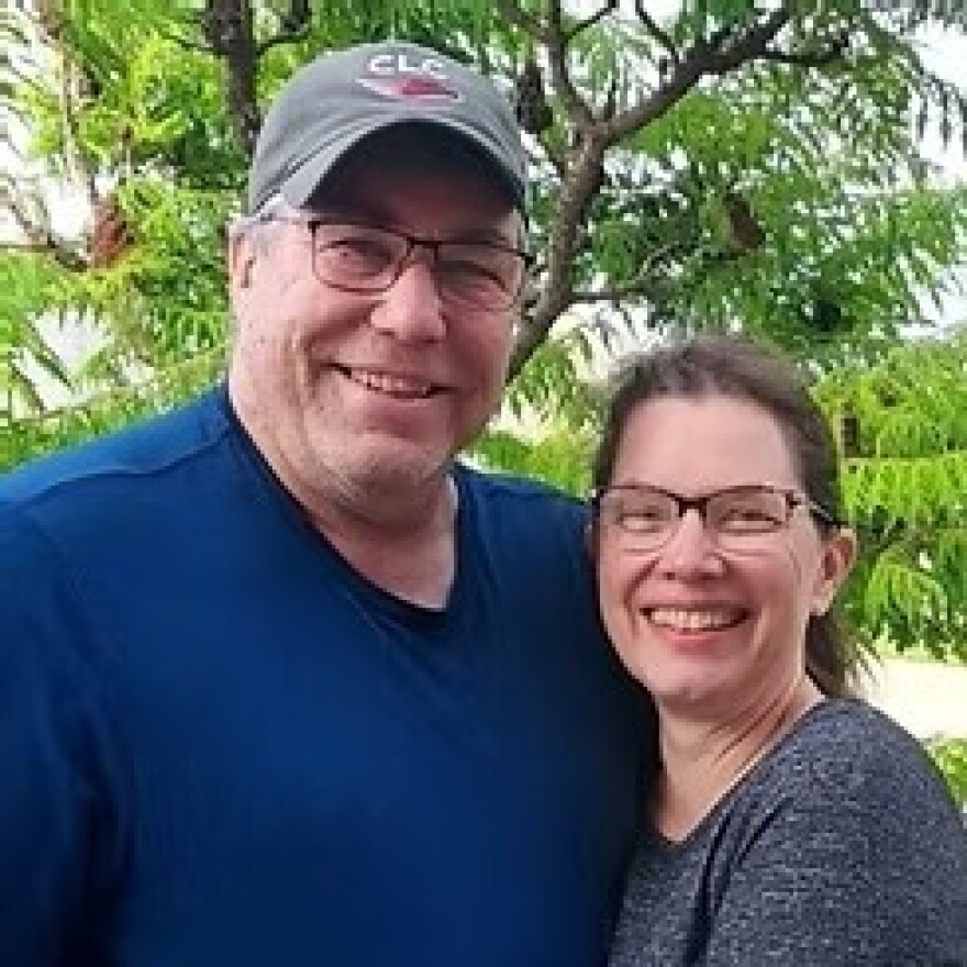 A husband and wife stand in front of a tree, smiling at the camera.