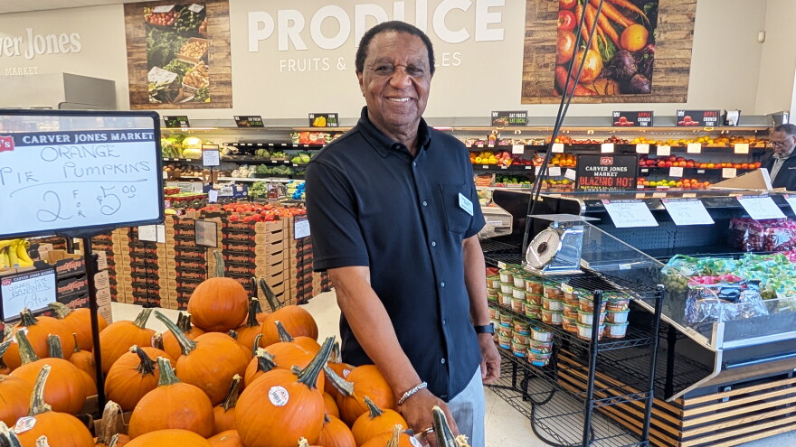 A man stands next to a bin of pumpkins that are priced two for $5. The man wears a black, button-down shirt and a name tag. The rest of a grocery store produce department is in the background.