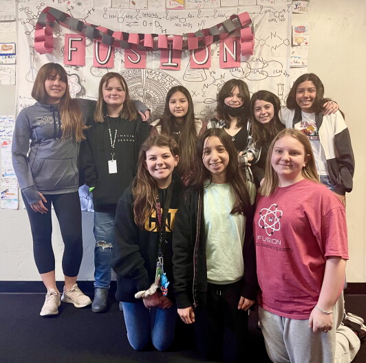 A teacher and eight students stand smiling together in front of a white and pink poster.