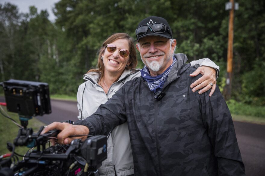 a woman and man stand next to each other and smile. they stand behind a film camera set-up