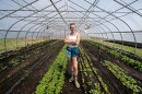 A person stands smiling with arms crossed inside a greenhouse, surrounded by rows of young plants on soil beds.