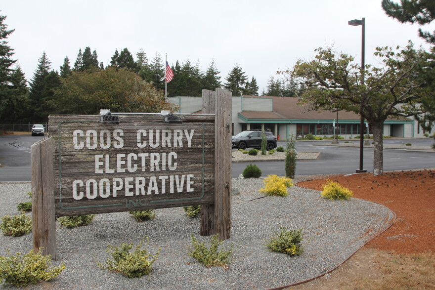 Wooden sign of the Coos Curry Electric Cooperative in a parking lot with municipal building and American flag in background.