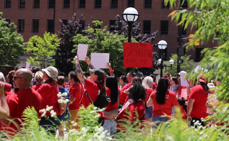 A medium shot captures a group of people at an outdoor protest or rally, mostly dressed in red shirts. Many individuals are holding up white signs, while a prominent red sign in the center-right reads "IT'S THE GASLIGHTING FOR ME" in black hand-drawn letters. The crowd is positioned in a plaza with green foliage in the foreground and a multi-story brick building in the background. A black lamppost with a white globe stands near the center of the frame. 