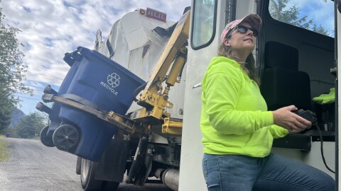 A woman in a bright neon hoodie operates a trash truck's mechanical arm, to pick up a recycling bin
