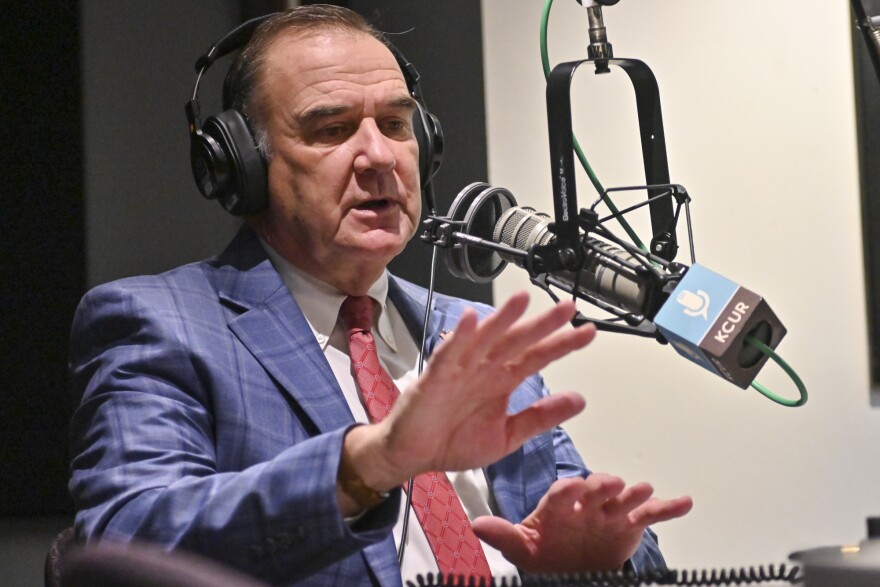 A man wearing a blue suit with red tie sits inside a radio studio. He is talking at a microphone and gesturing with both hands.