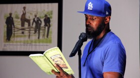 Ronnie Amiyn, wearing a blue shirt and blue Cardinals baseball hat, stands at a microphone during a poetry reading. He reads from a book written by Bobby Bostic. 