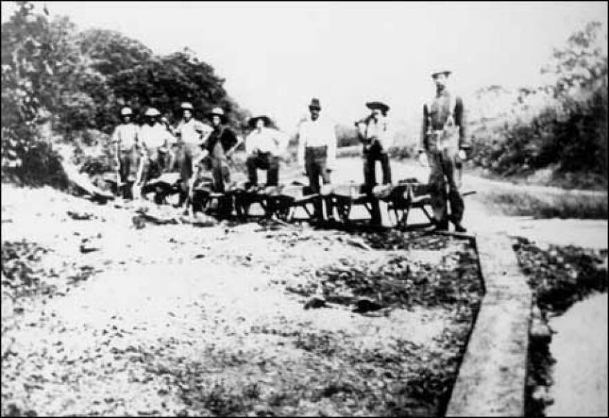 A black and white photograph shows a group of men  standing on a dirt path next to a wooden walkway. The men appear to be laborers, some holding what look like wheelbarrows, suggesting they are working on a construction project