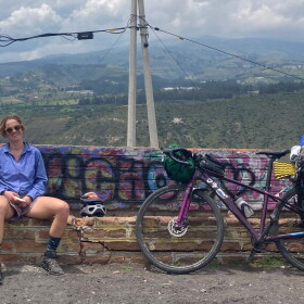 Smiling woman in shorts sitting on bench with graffiti. Two biked leaning against the bench filled with rucksacks. Mountains in background