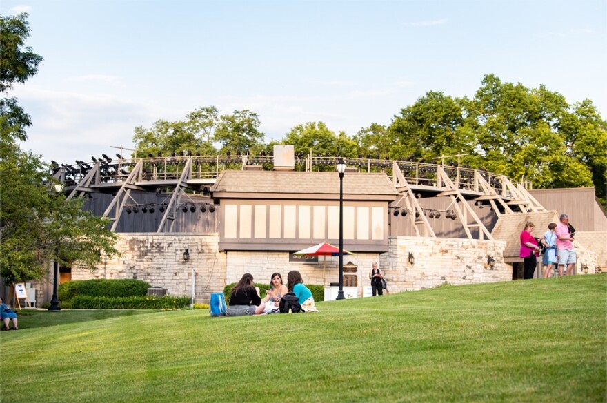 A green, grassy knoll with people picnicking foregrounds the Ewing Cultural Center's round brick and wooden theater.