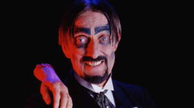 Man dressed in a costume with black suit and a severed hand on his shoulder 