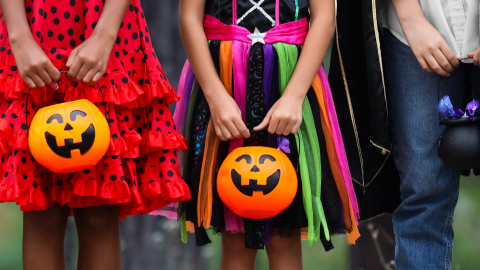 Three children dressed in colorful Halloween costumes hold orange jack-o'-lantern candy buckets, ready for trick-or-treating.